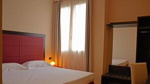 Habitació d'hotel amb un llit gran, una taula de nit amb una làmpada, una cadira i un armari.