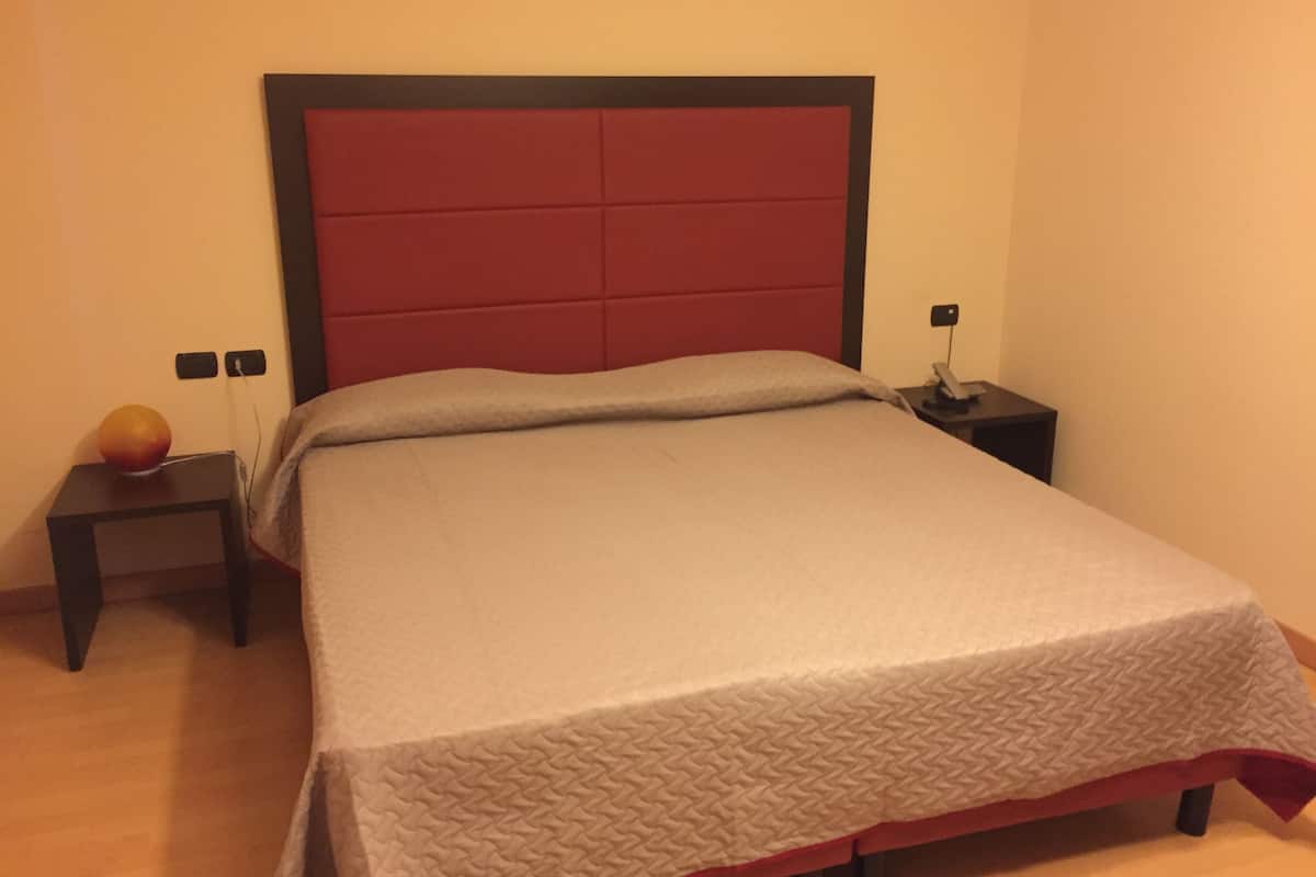 Una cama bien hecha con cabecera roja, una mesita de noche con lámpara y un enchufe de pared.