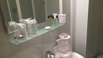 Une salle de bain avec une étagère en verre où sont rangés les articles de toilette, un lavabo blanc et une serviette enroulée posée sur le sol.