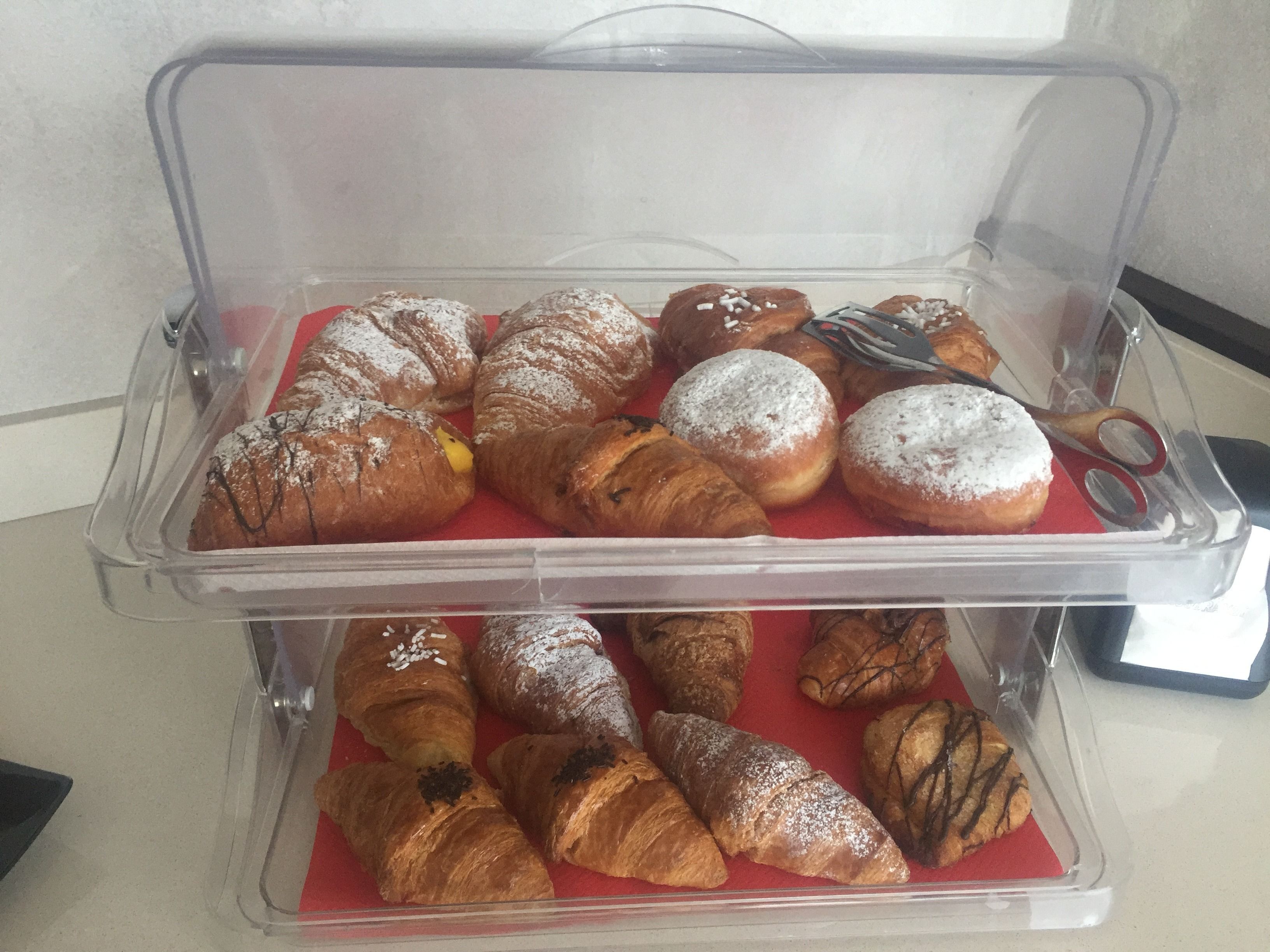 A clear plastic container with a variety of pastries, including croissants and danishes, some dusted with powdered sugar.