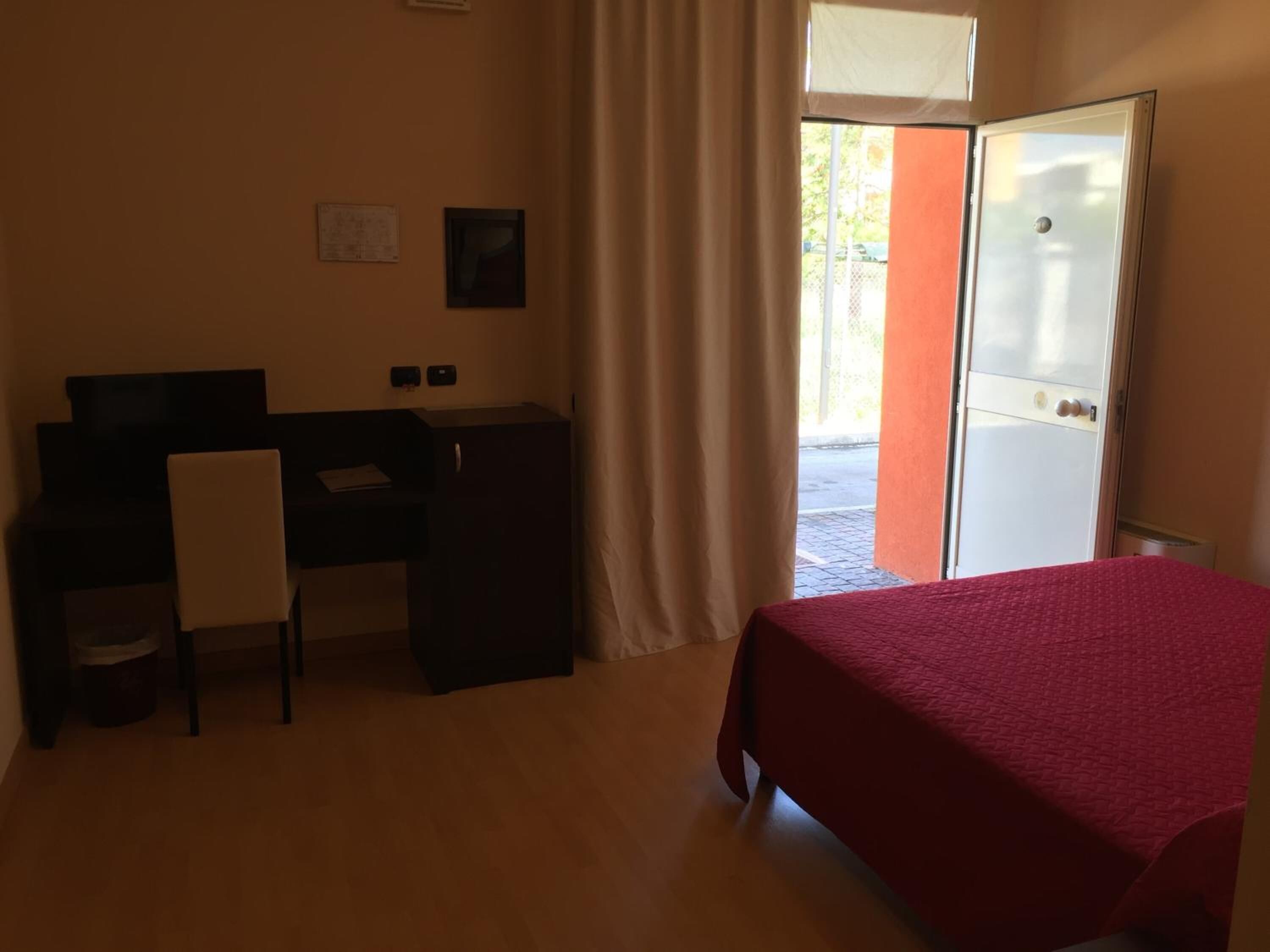 Una habitación con cama, escritorio y silla. Hay una ventana con cortinas y una puerta que da al exterior.