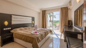 A hotel room with a large bed, a desk, a TV, and a balcony with a view of palm trees. - Cavo D'oro Hotel (Kos)