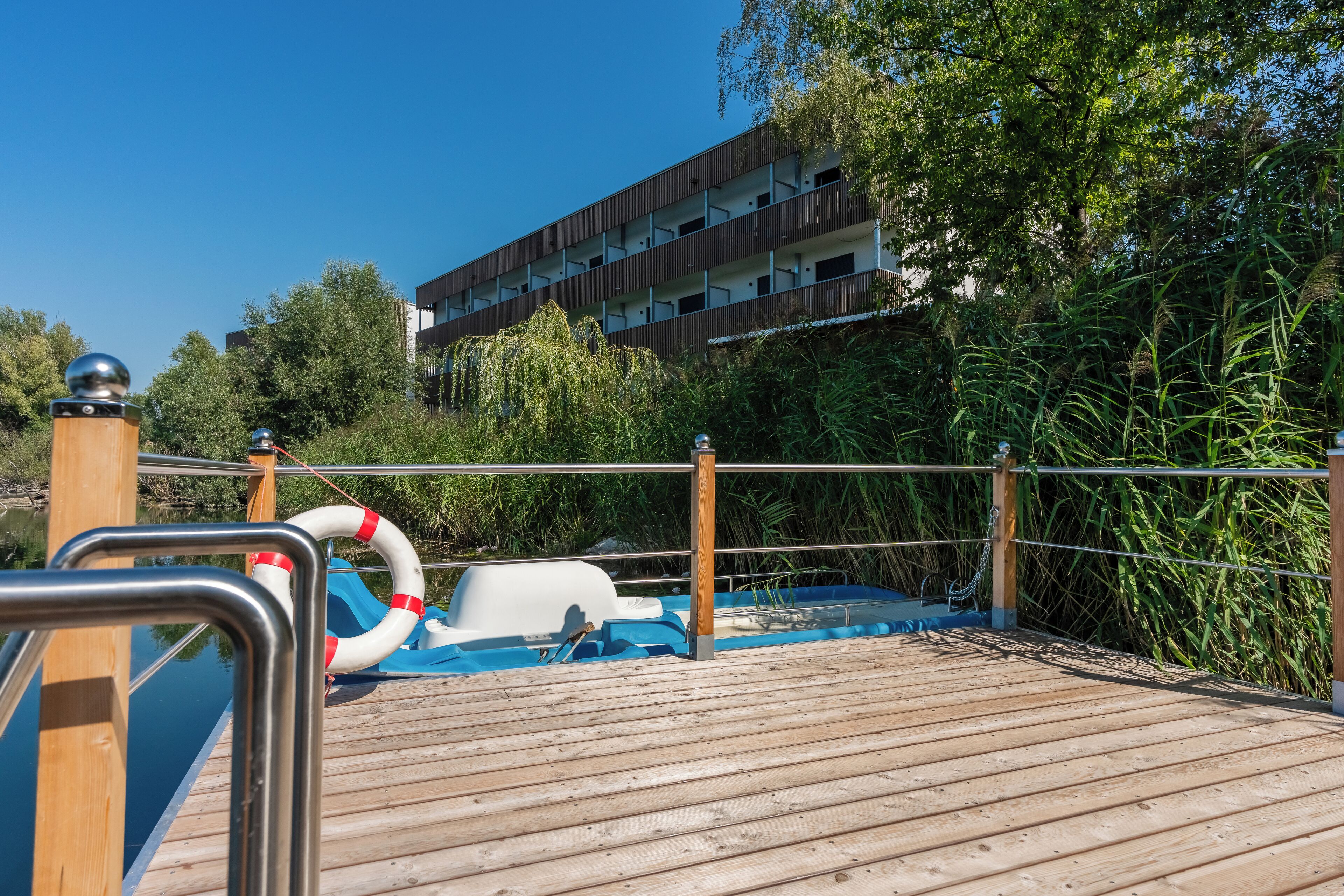 A wooden deck with a pool, surrounded by greenery and a building in the background.
