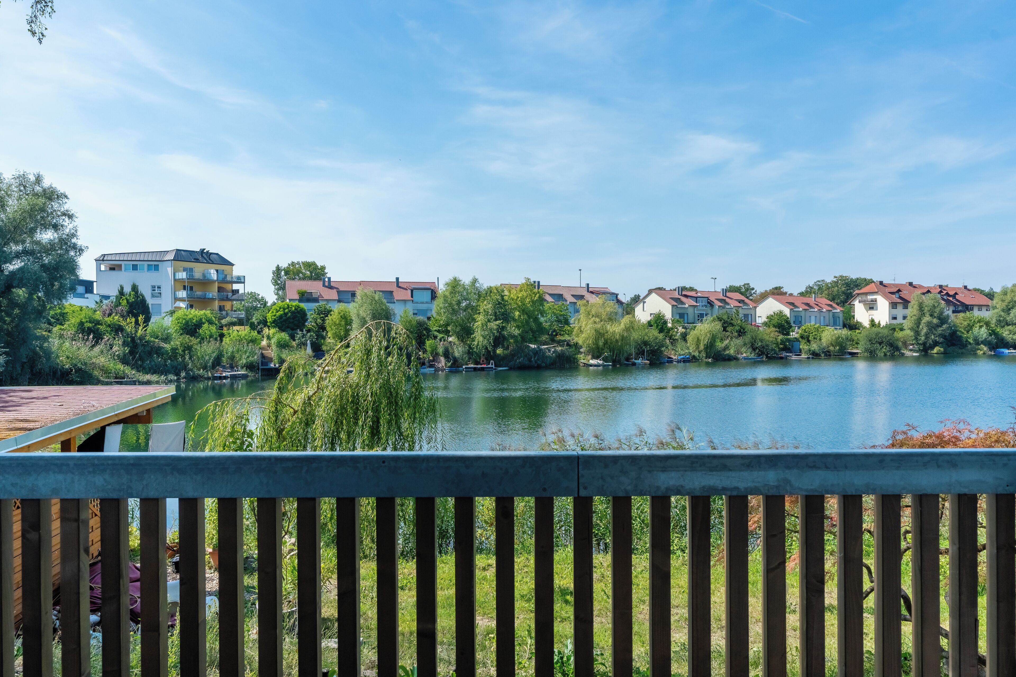 A lakeside view with residential buildings, a wooden deck, and lush greenery.