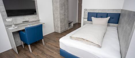 A modern hotel room with a large bed, a desk with a flat-screen TV, a wardrobe, and a blue chair.