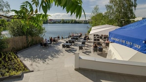 An outdoor patio area with tables and chairs, overlooking a body of water, and a blue tent with text. - Seehotel Forst (Forst)