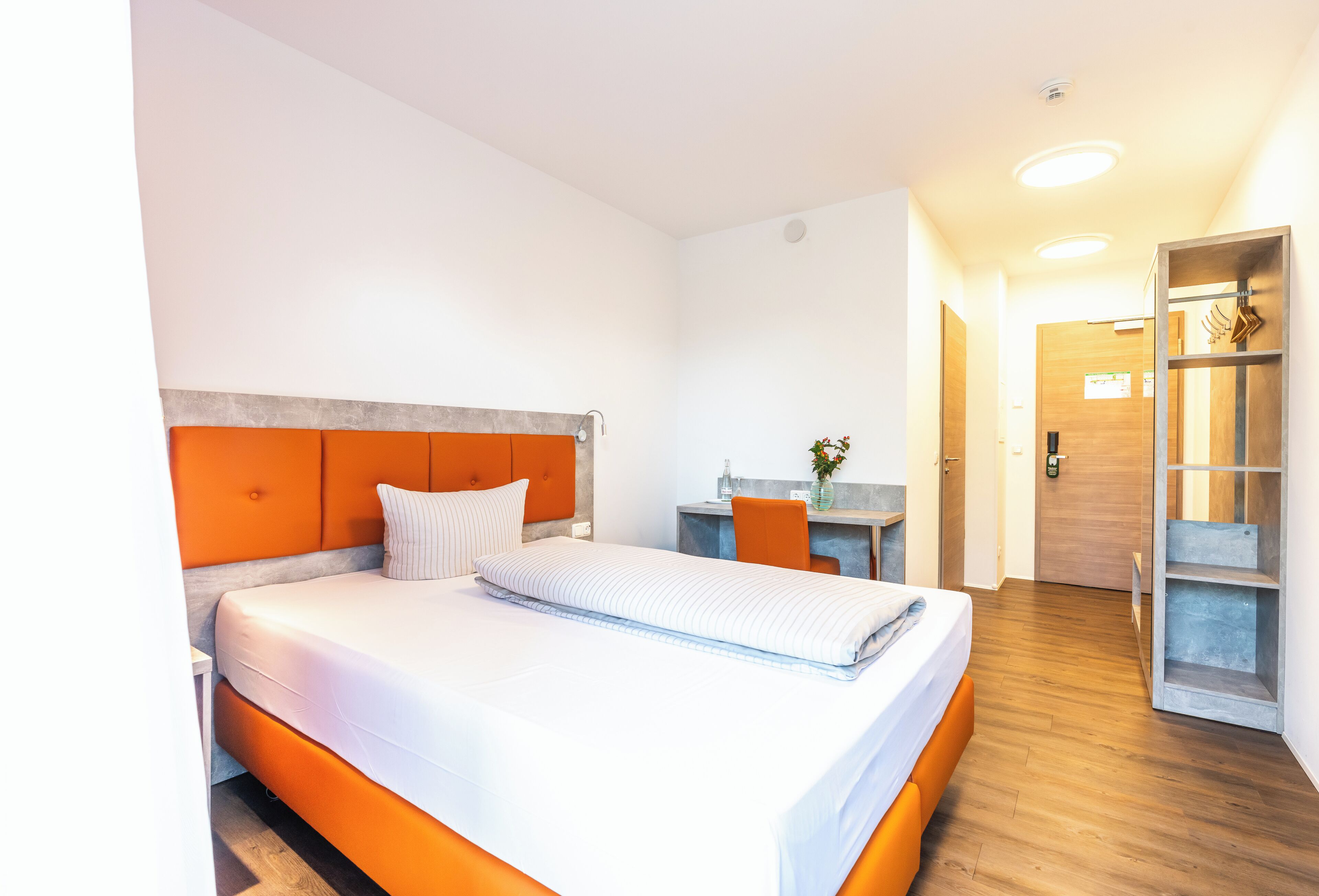 Habitación de hotel moderna con una cama grande, suelo de madera, un escritorio pequeño y un baño a la vista al fondo.