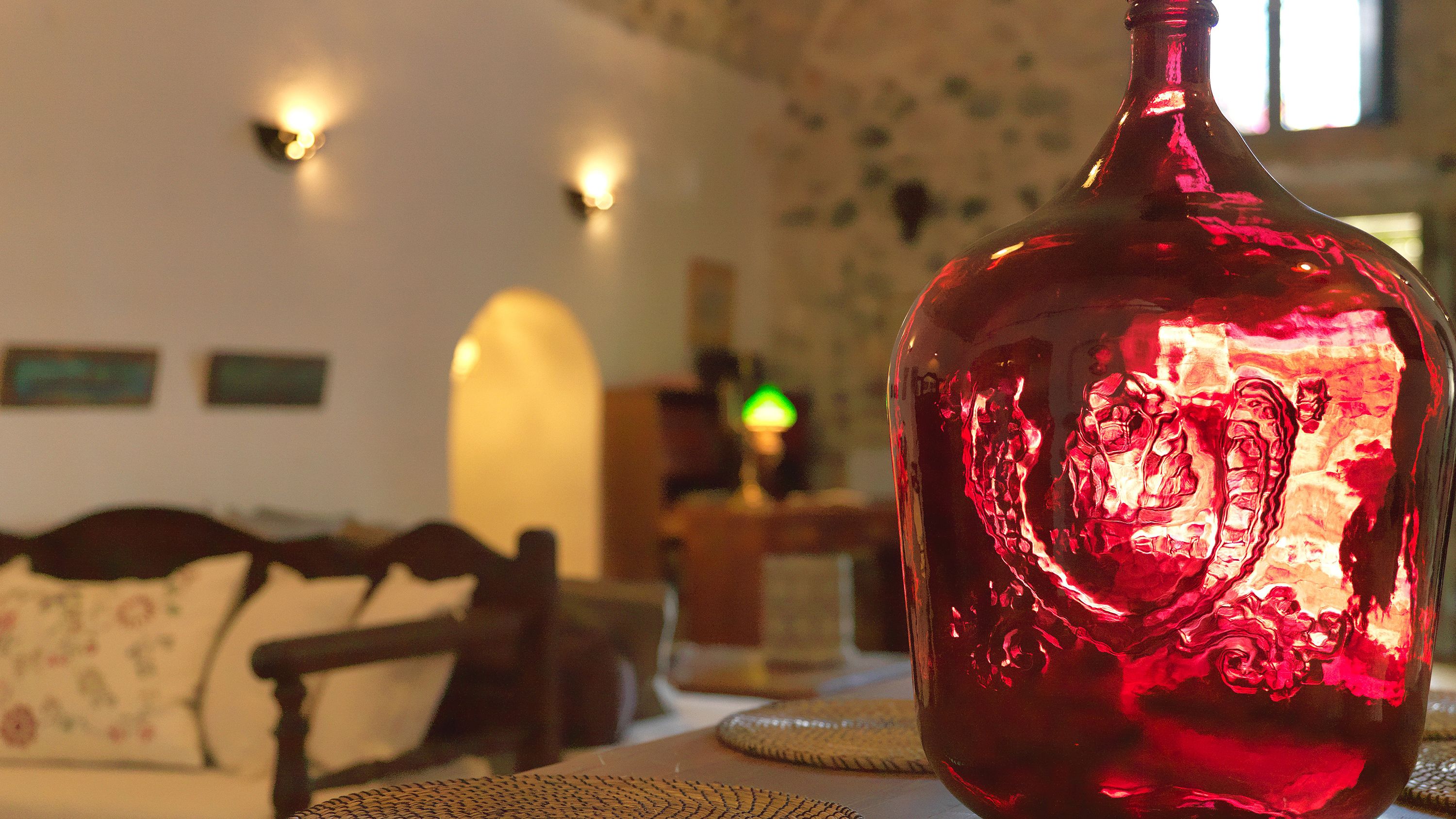 Uma garrafa de vidro vermelha com design texturizado sobre uma mesa em um quarto com sofá e luminárias na parede.