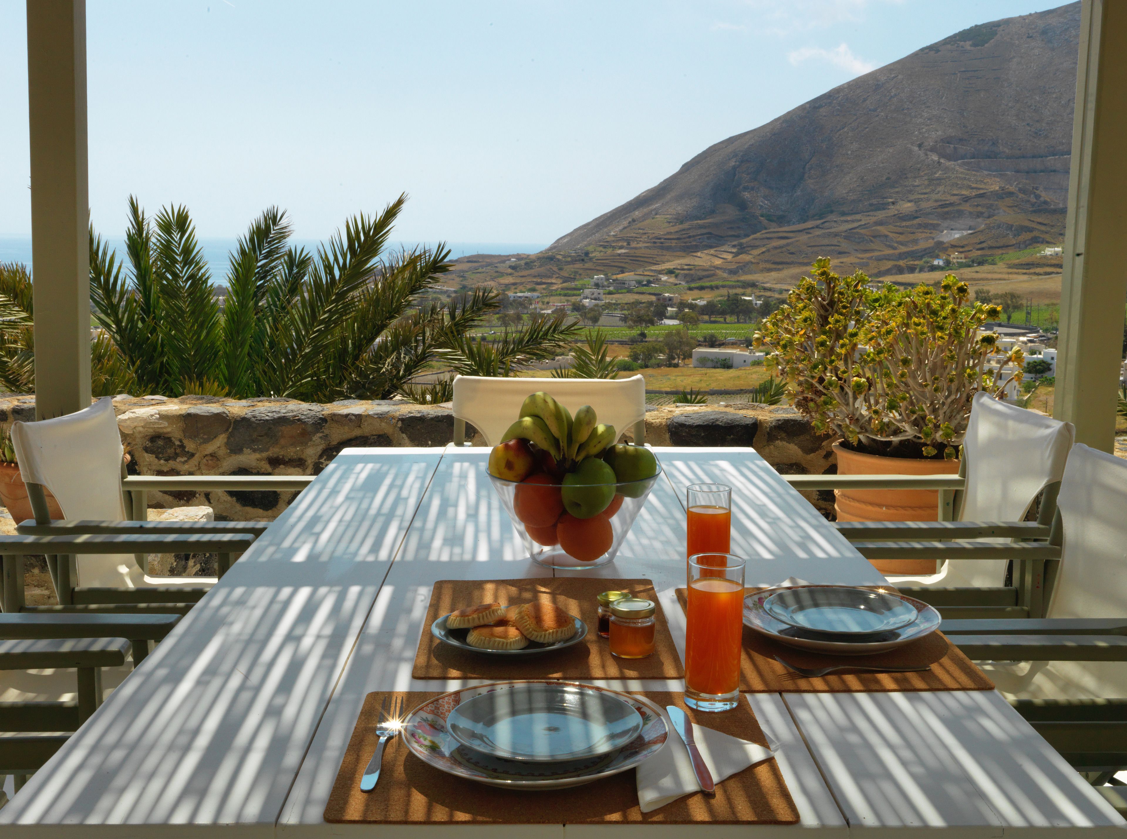 A table set with plates, cutlery, a bowl of fruit, and a glass of orange juice, overlooking a scenic landscape with mountains and a village.