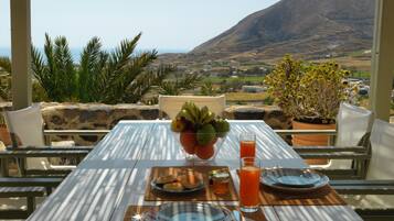 A table set with plates, cutlery, a bowl of fruit, and a glass of orange juice, overlooking a scenic landscape with mountains and a village.