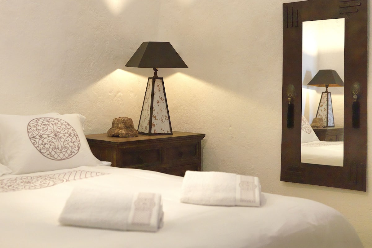 A neatly made bed with white linens and decorative pillows, a bedside table with a lamp, and a mirror reflecting the lamp.
