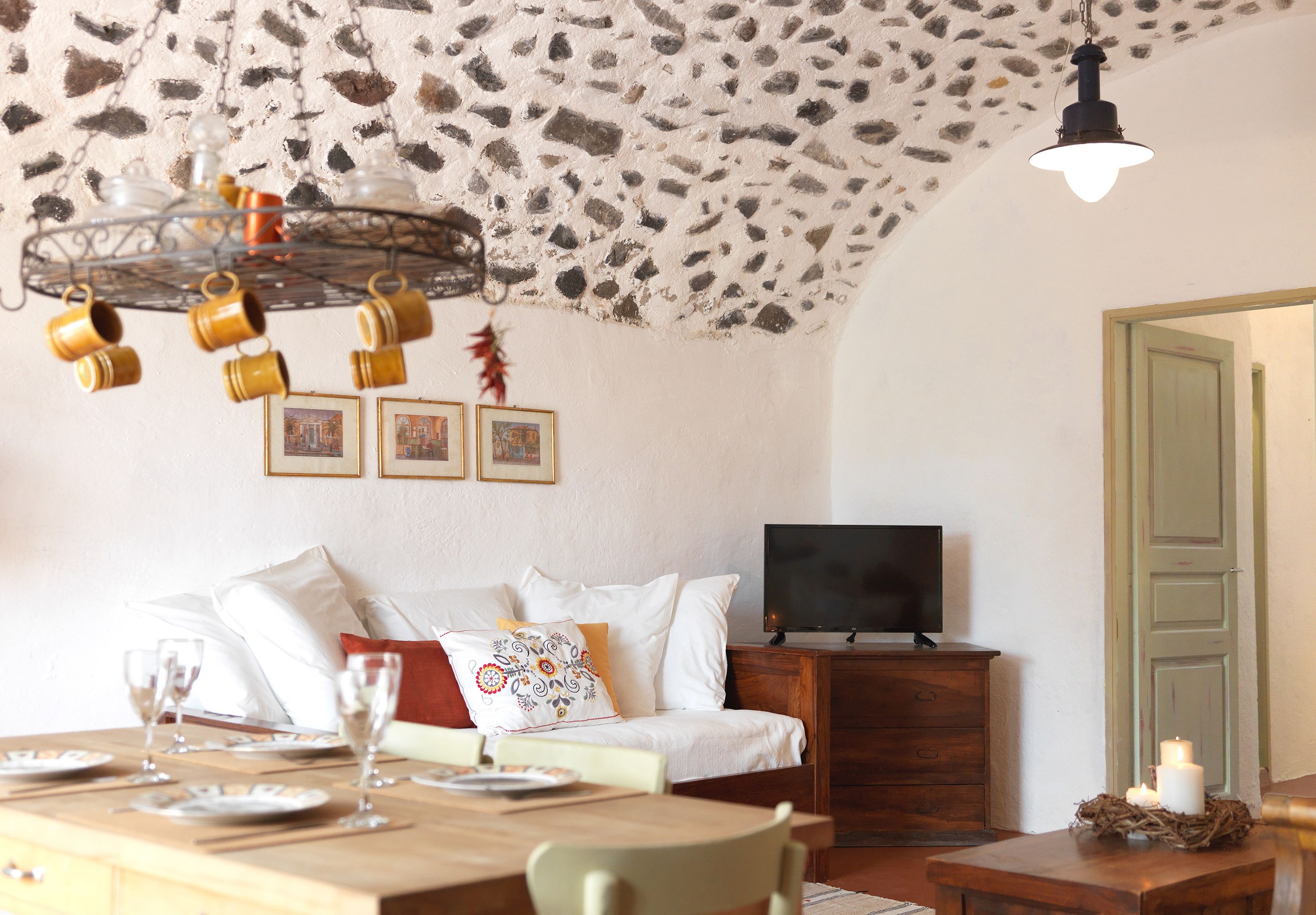 Una sala de estar acogedora con techo de piedra, una mesa de comedor de madera servida con platos y vasos, un sofá con cojines y un televisor sobre un mueble de madera.