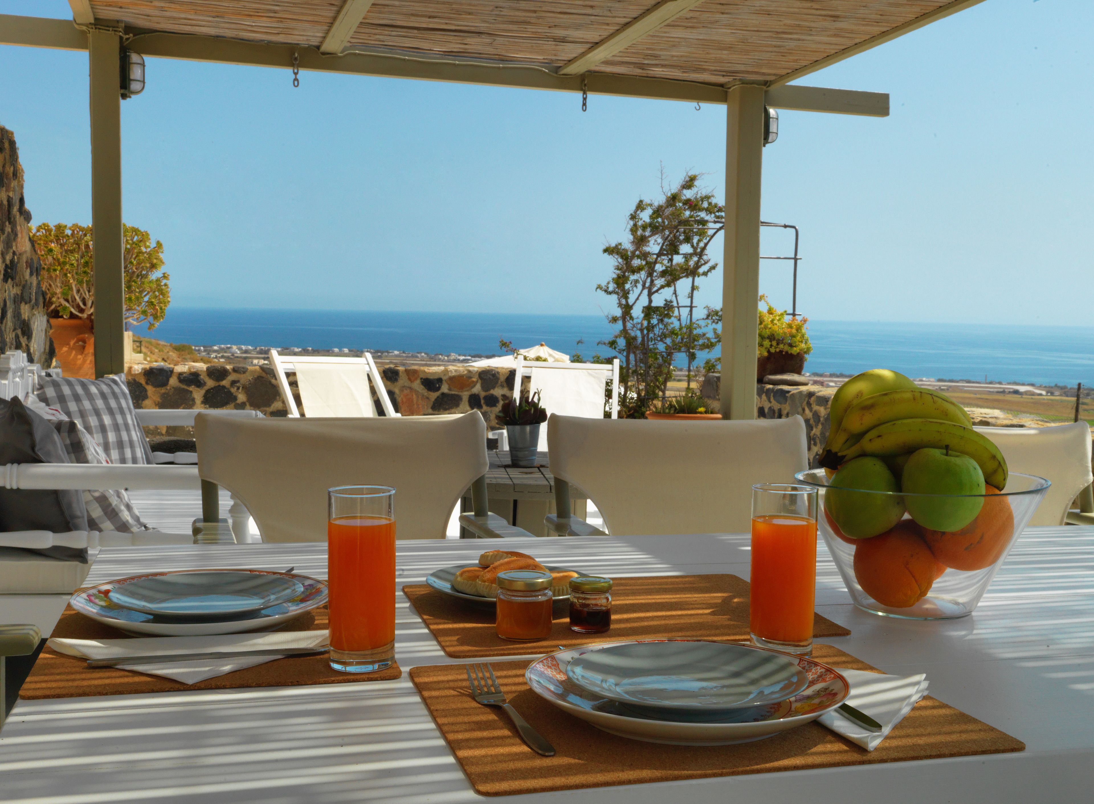 A table set for breakfast with a view of the sea, including plates, glasses, and a bowl of fruit.