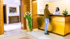 Zona de recepción con un mostrador de madera, una recepcionista y un huésped de pie.