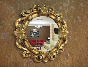 A mirror with an ornate gold frame reflecting a room with a red sofa, a white armchair, and a chandelier.