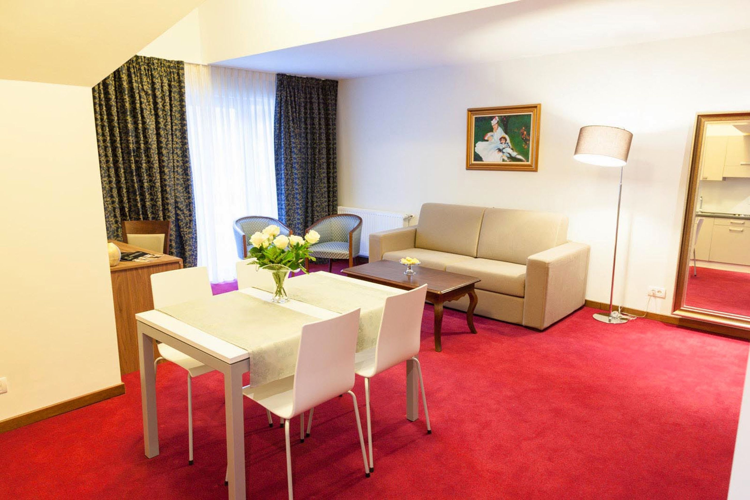 A hotel room with a dining table, two chairs, a sofa, a floor lamp, and a painting on the wall.
