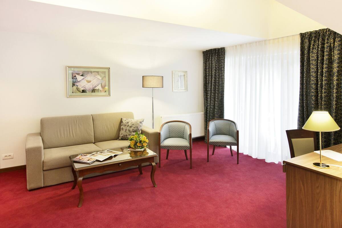 Habitació d'hotel amb un sofà, dues butaques, una taula de centre amb revistes, un escriptori amb una làmpada i una finestra amb cortines.