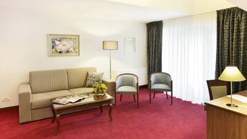Habitación de hotel con sofá, dos sillones, una mesita de centro con revistas, un escritorio con lámpara y una ventana con cortinas.