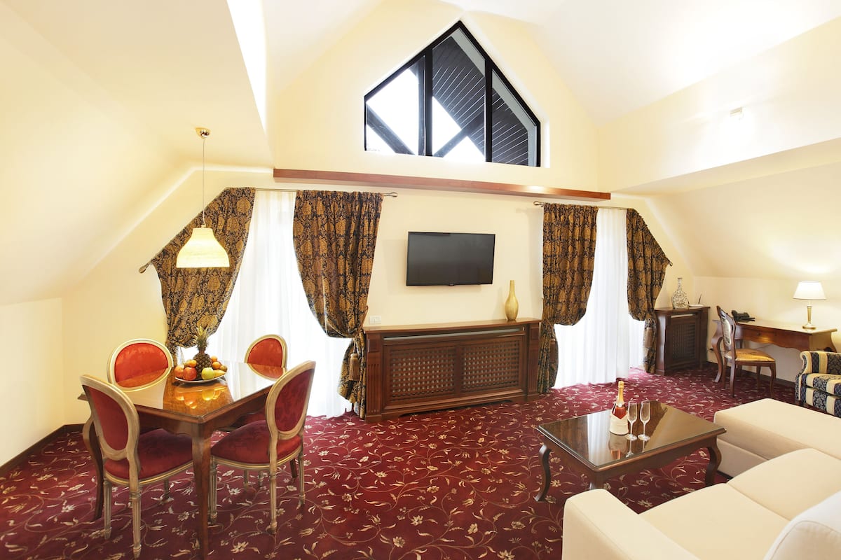 Habitació d'hotel amb zona de menjador, saló amb sofà i butaques, televisió penjada a la paret i una finestra gran amb cortines.