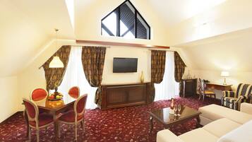 A hotel room with a dining area, a living area with a sofa and armchairs, a TV mounted on the wall, and a large window with curtains.