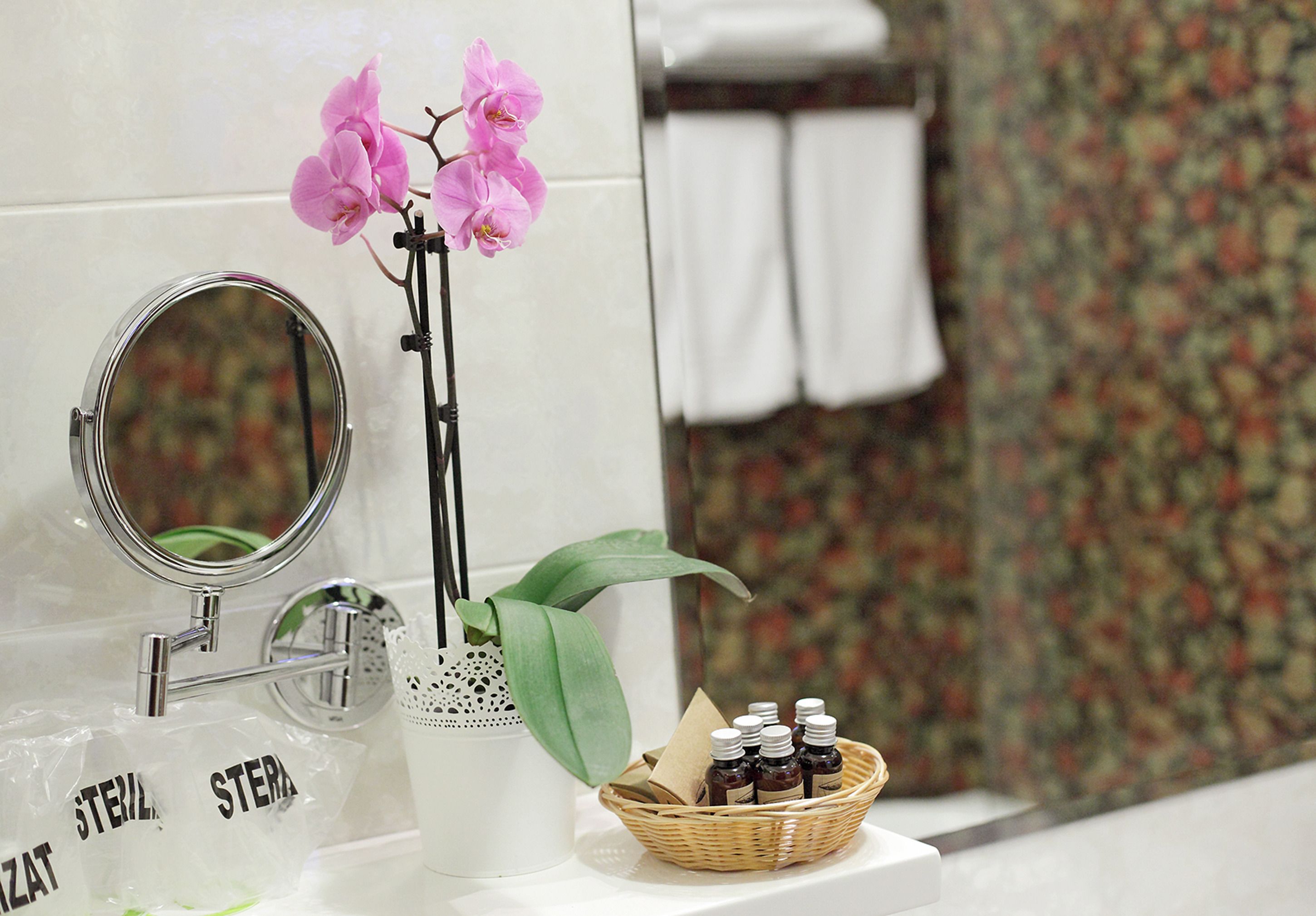 Un tocador de baño con espejo, una orquídea en maceta, una cesta de mimbre con artículos de aseo y un recipiente de vidrio decorativo.