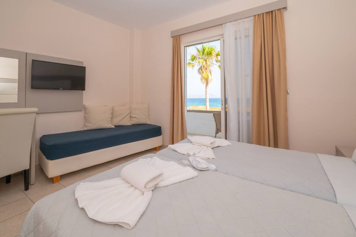 Habitación de hotel con cama, sofá, televisión y balcón con vista a una palmera.