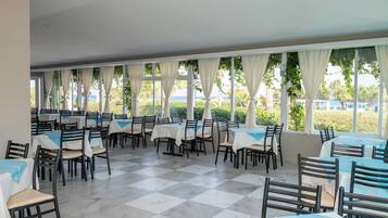 A dining area with tables and chairs, large windows with curtains, and a view of greenery and buildings.