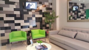 A modern living room with a checkered wall, two green armchairs, a grey sofa, a white coffee table with books, and a TV mounted on the wall. - Portofino Hotel (Teresina)