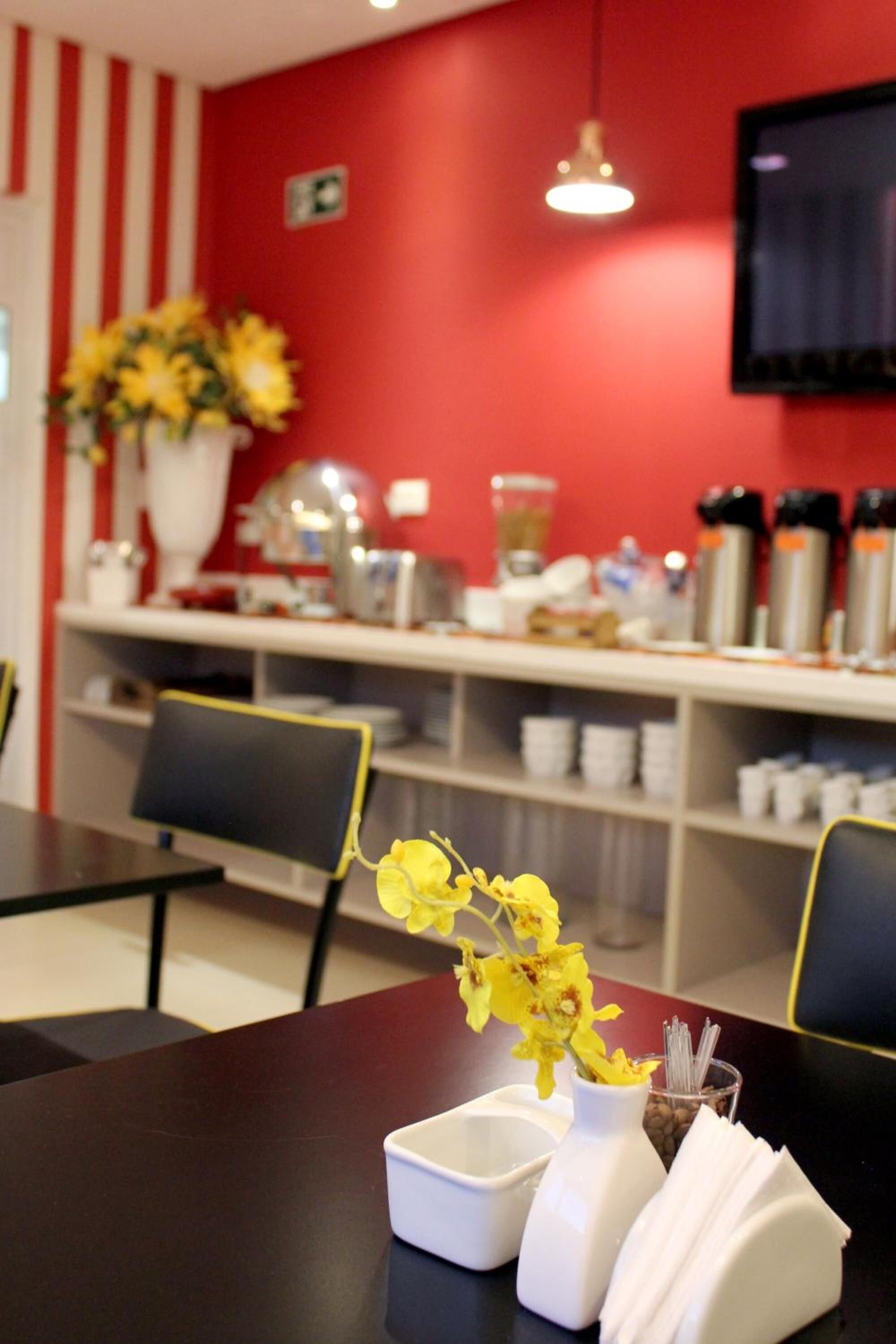 A restaurant interior with a red wall, a white vase with yellow flowers, a coffee machine, and a TV mounted on the wall.