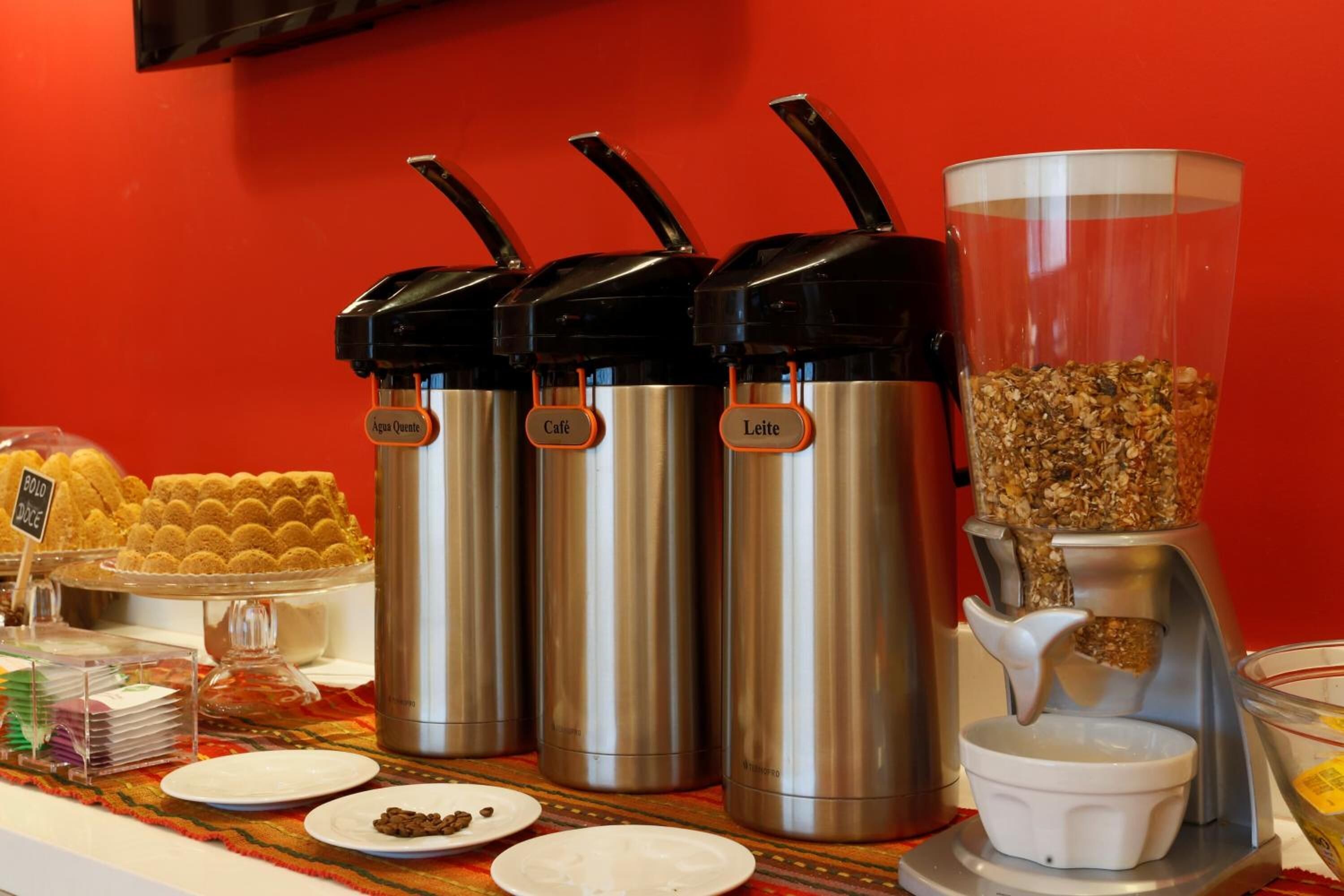 A buffet setup with three stainless steel dispensers labeled 'Aqua Quente', 'Café', and 'Leite', and a coffee dispenser with a granola container.