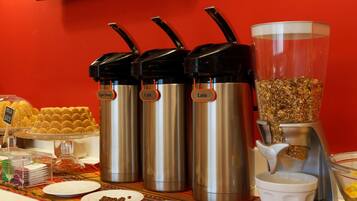 A buffet setup with three stainless steel dispensers labeled 'Aqua Quente', 'Café', and 'Leite', and a coffee dispenser with a granola container.