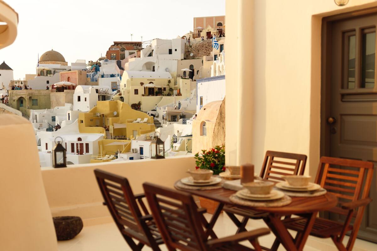 Un balcón con vistas a edificios blancos, una mesa con platos y una vela, y sillas de madera.