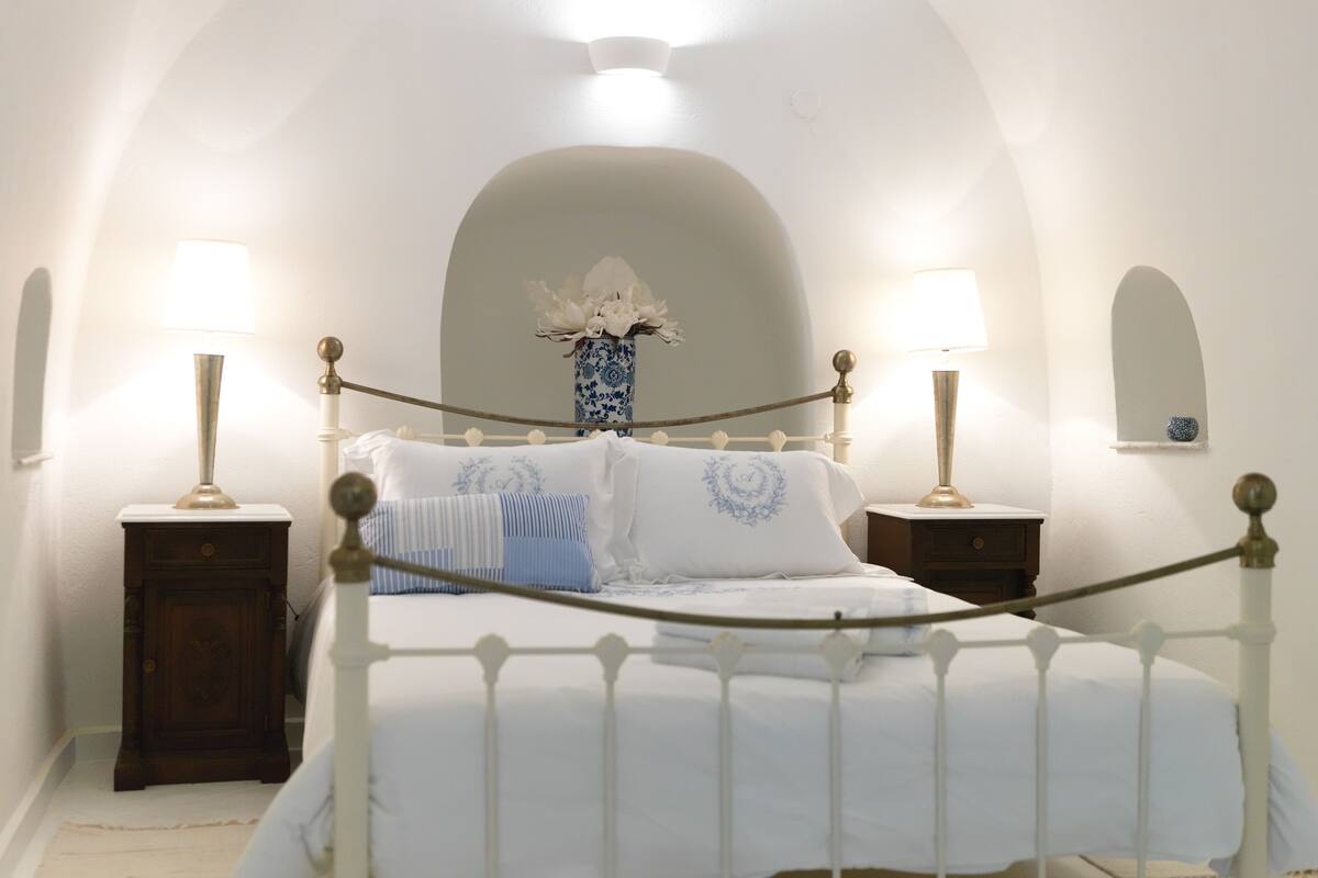 Un dormitorio con una cama blanca, dos mesitas de noche con lámparas, un jarrón con flores y un techo abovedado blanco.