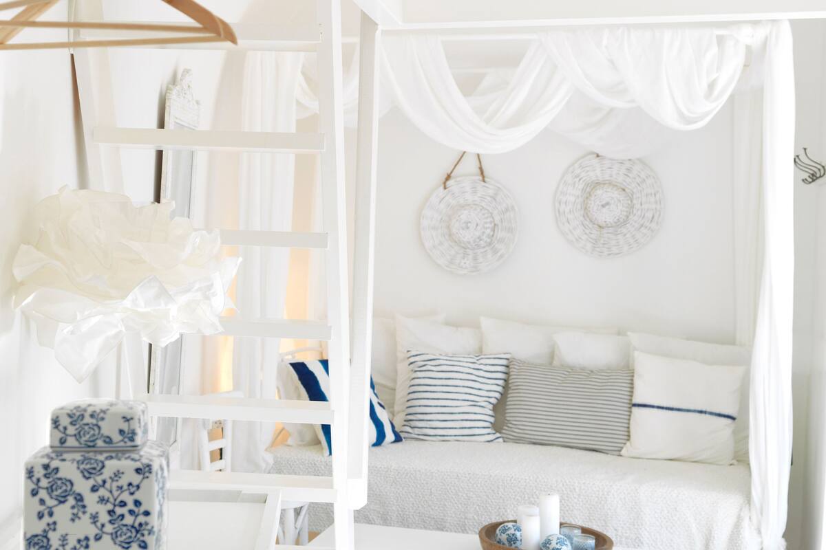 Una habitación blanca con una cama, una silla blanca, un estante blanco y una mesa blanca.