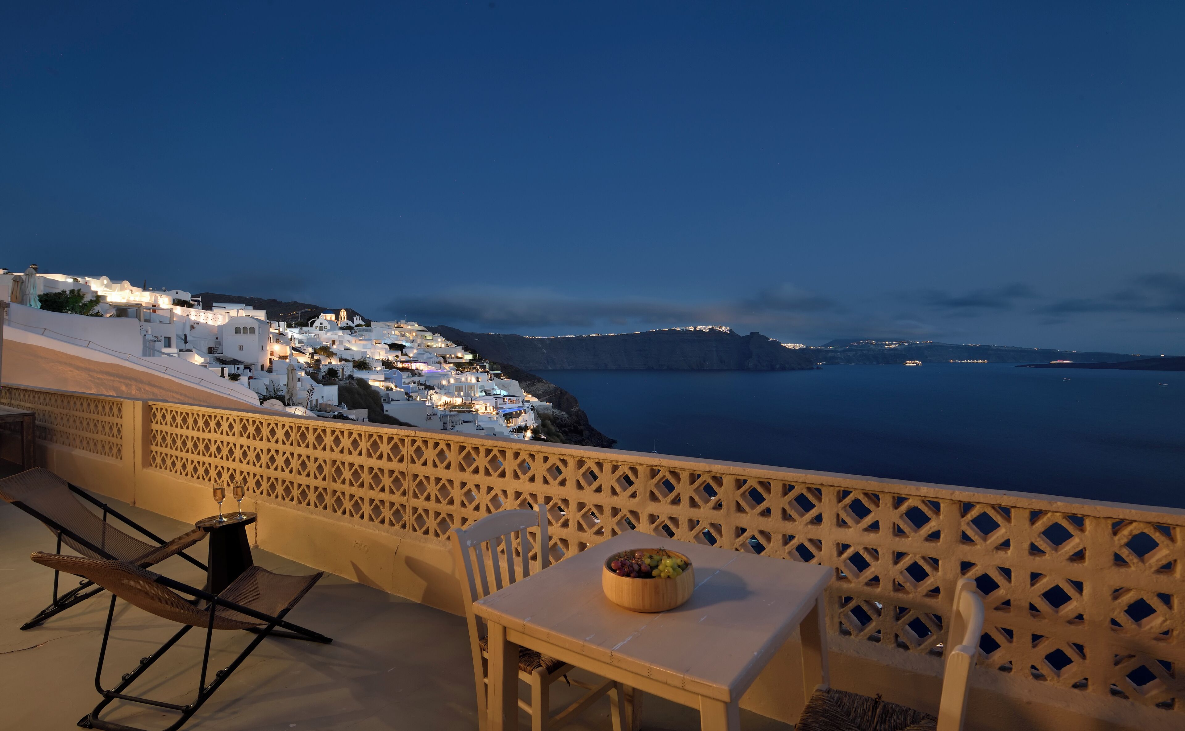 A balcony with a table and chairs, overlooking a coastal town at dusk.