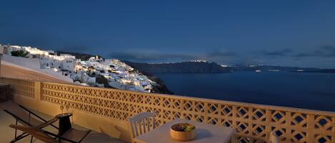 A balcony with a table and chairs, overlooking a coastal town at dusk.