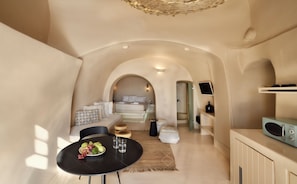 A compact, well-lit room with a bed, a dining table with fruits, and a kitchenette. - OIA Unique Homes by K&K (Santorini)