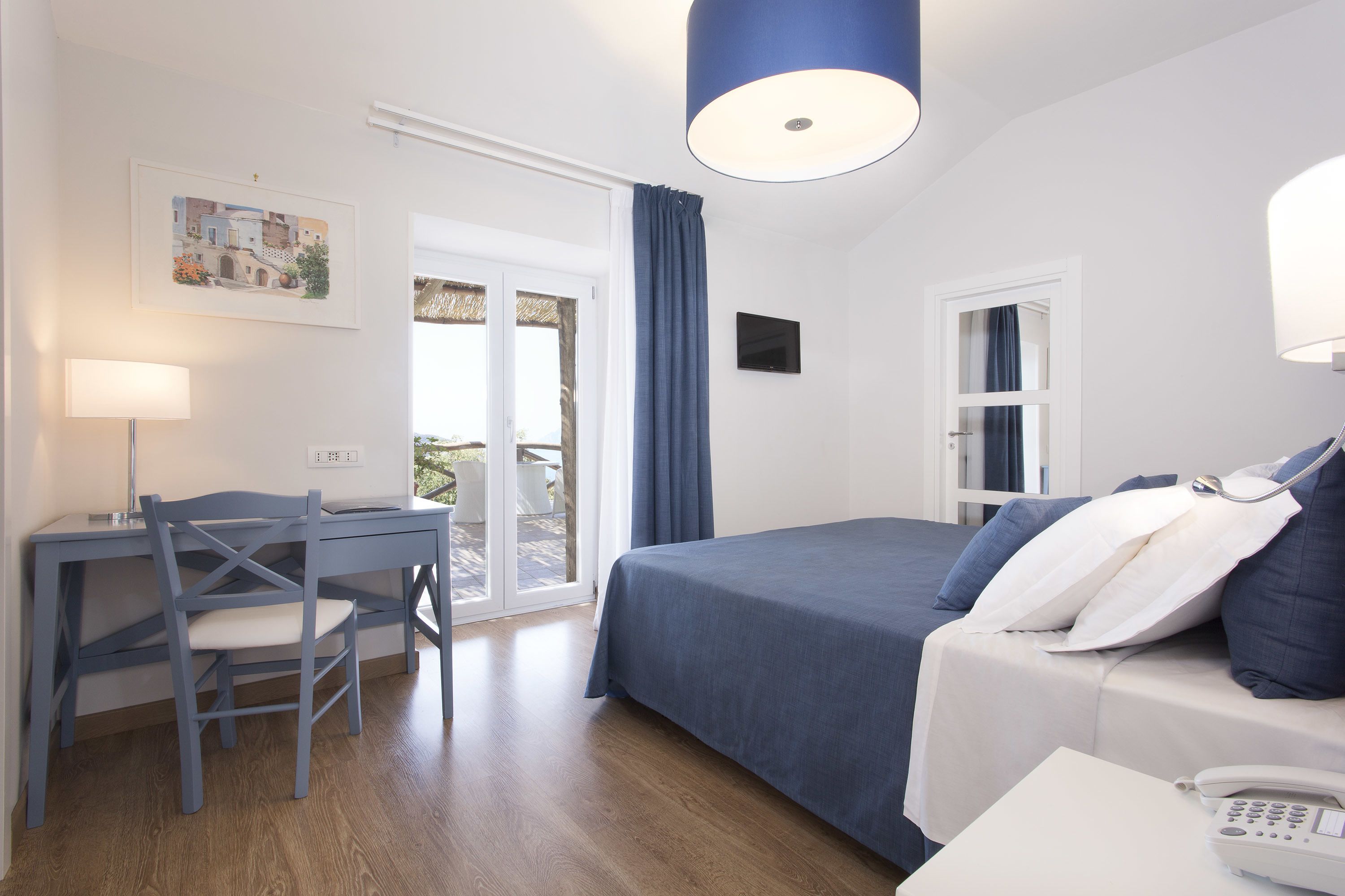 Ein Schlafzimmer mit einem Bett, einem Schreibtisch und einem Stuhl. Ein Fenster mit Vorhängen, eine Tür und ein kleiner Tisch mit einem Telefon sind ebenfalls vorhanden.