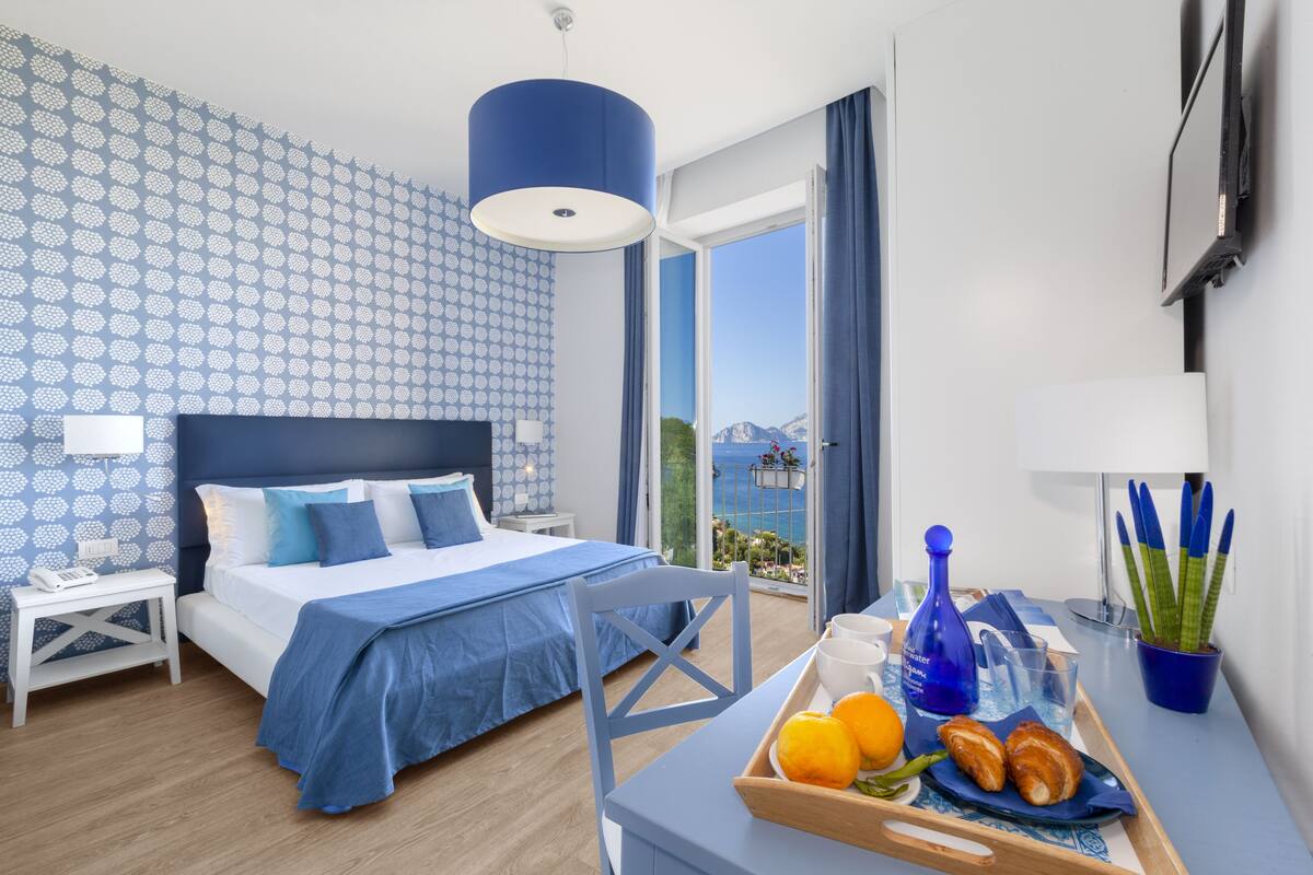 A hotel room with a bed, a table with breakfast items, a potted plant, and a view of the sea.