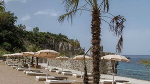 Un espacio de descanso junto a la playa con jardineras de madera, sombrillas color beige y muros de piedra.