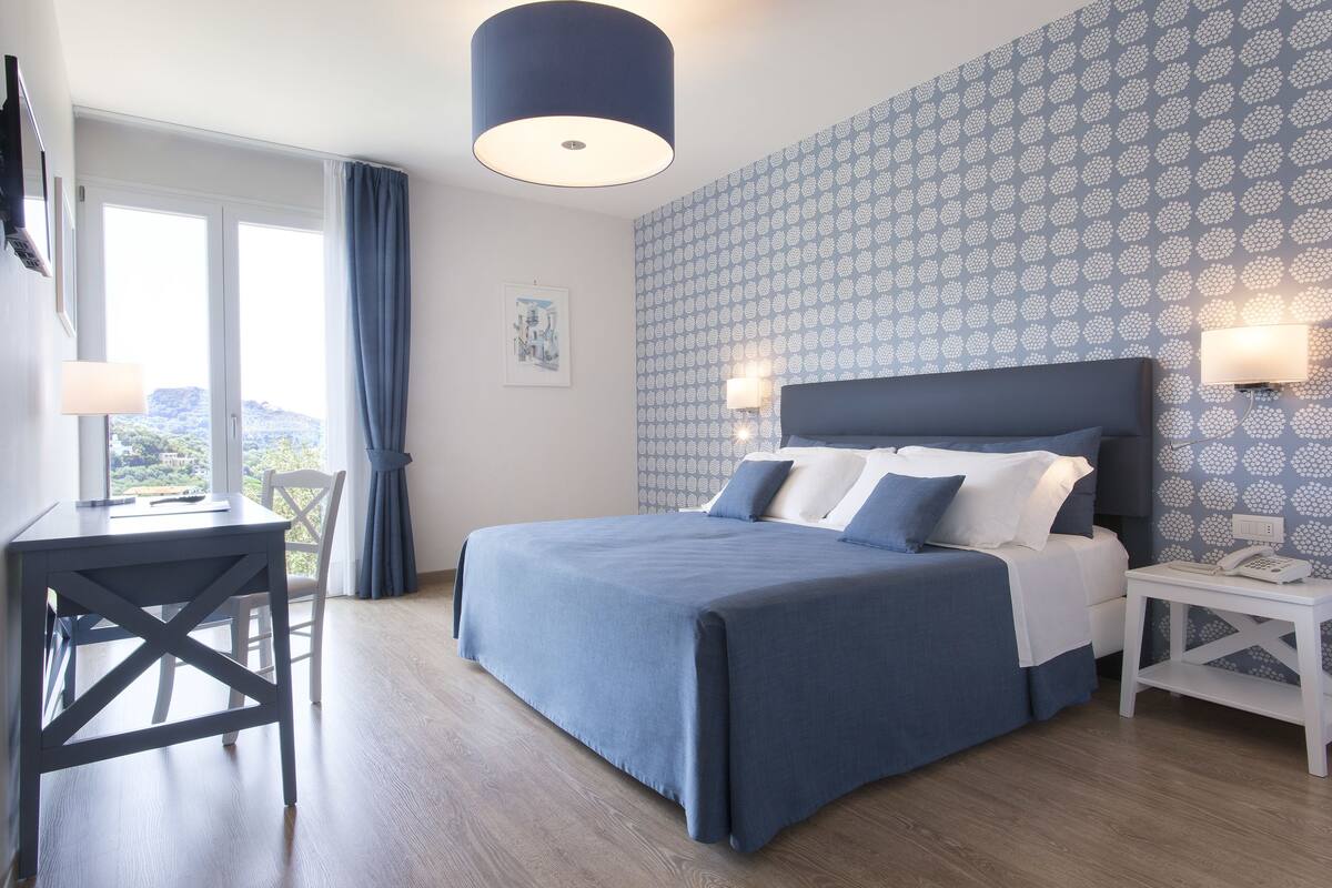 A bedroom with a bed, a desk, and a chair. There is a window with curtains, a wall with a patterned wallpaper, and a ceiling light.