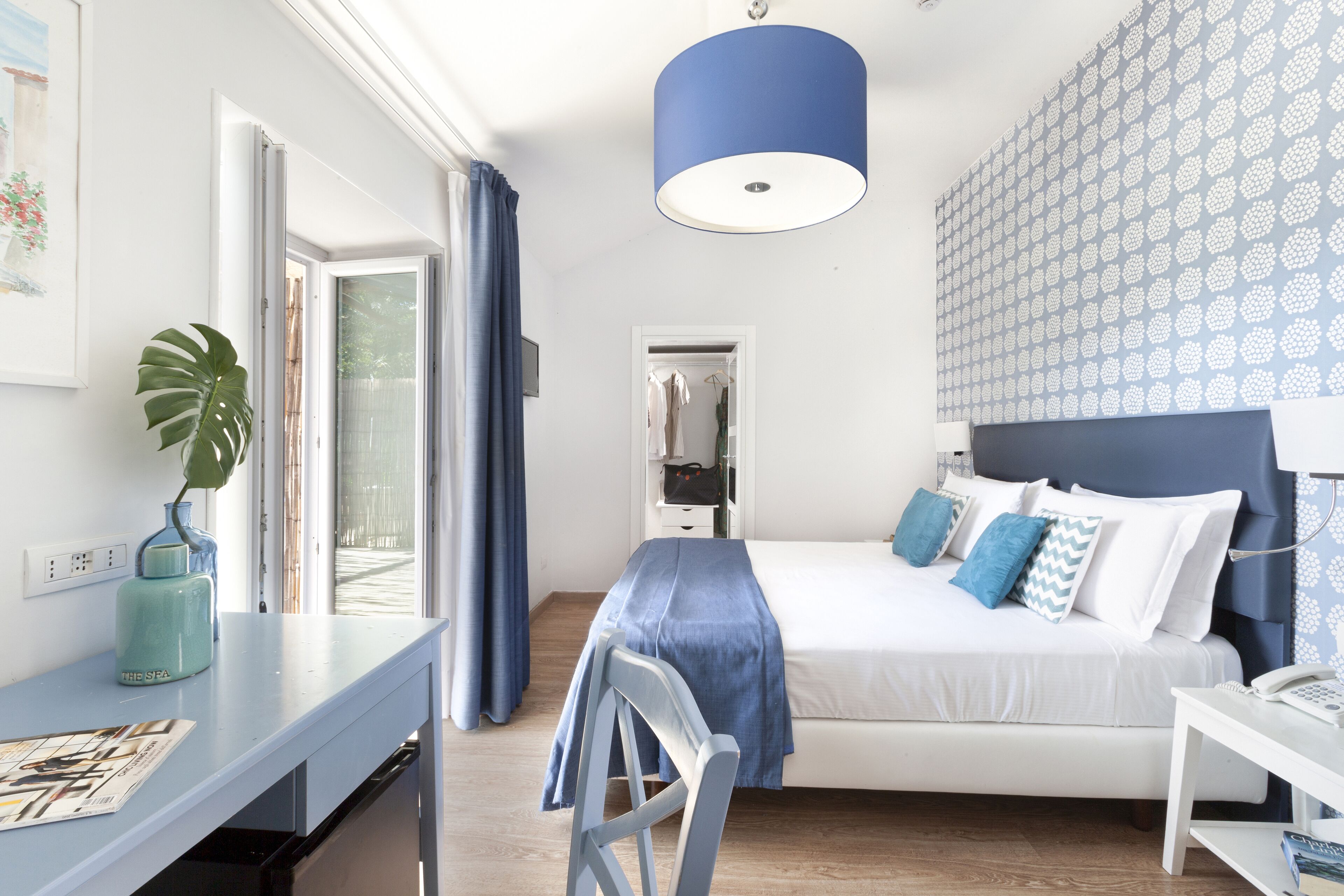 A bedroom with a bed, a desk, and a chair. There is a window with curtains, a plant, and a wall with a pattern.