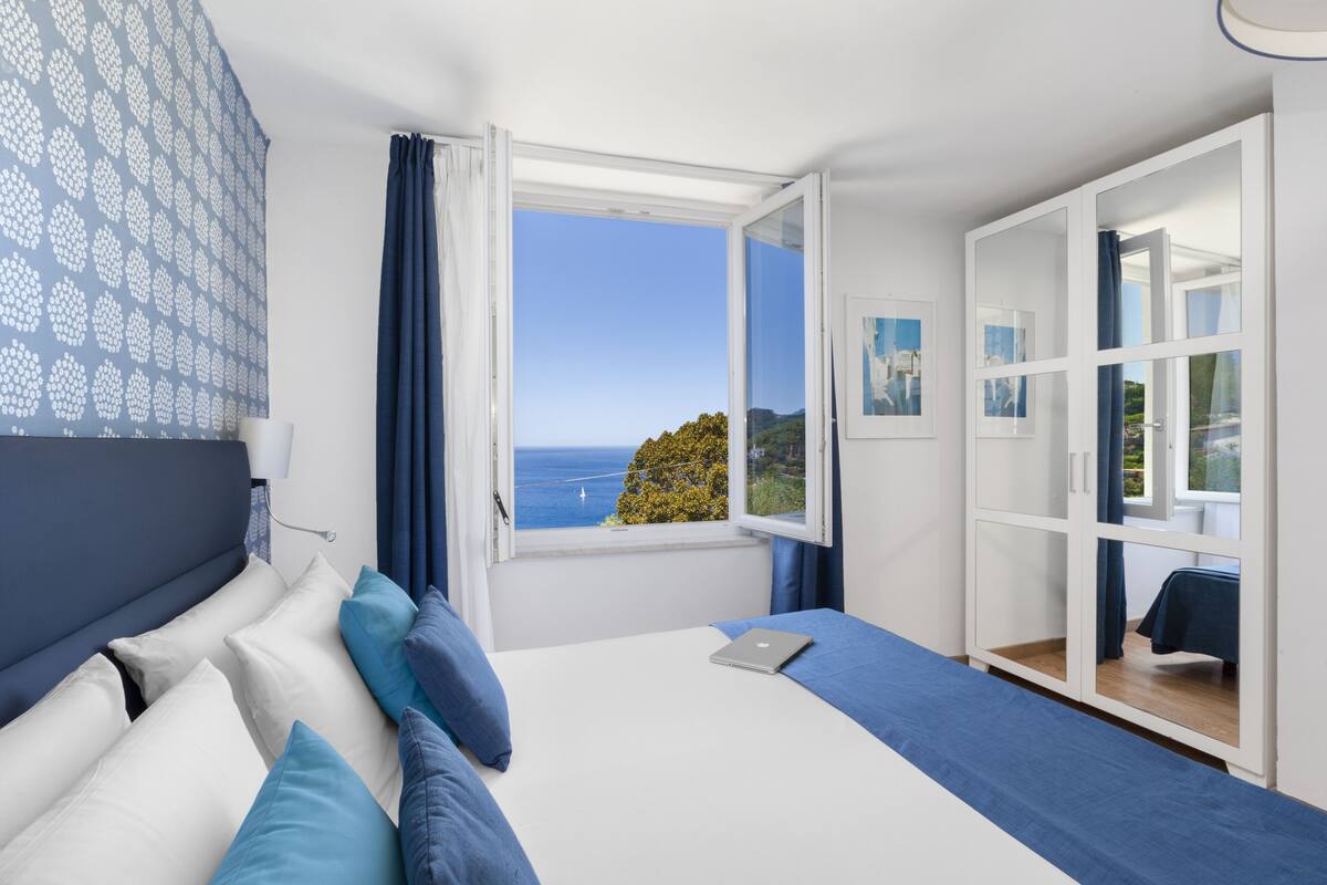 A bedroom with a bed, a laptop, a window with a view of the sea, and a wardrobe.