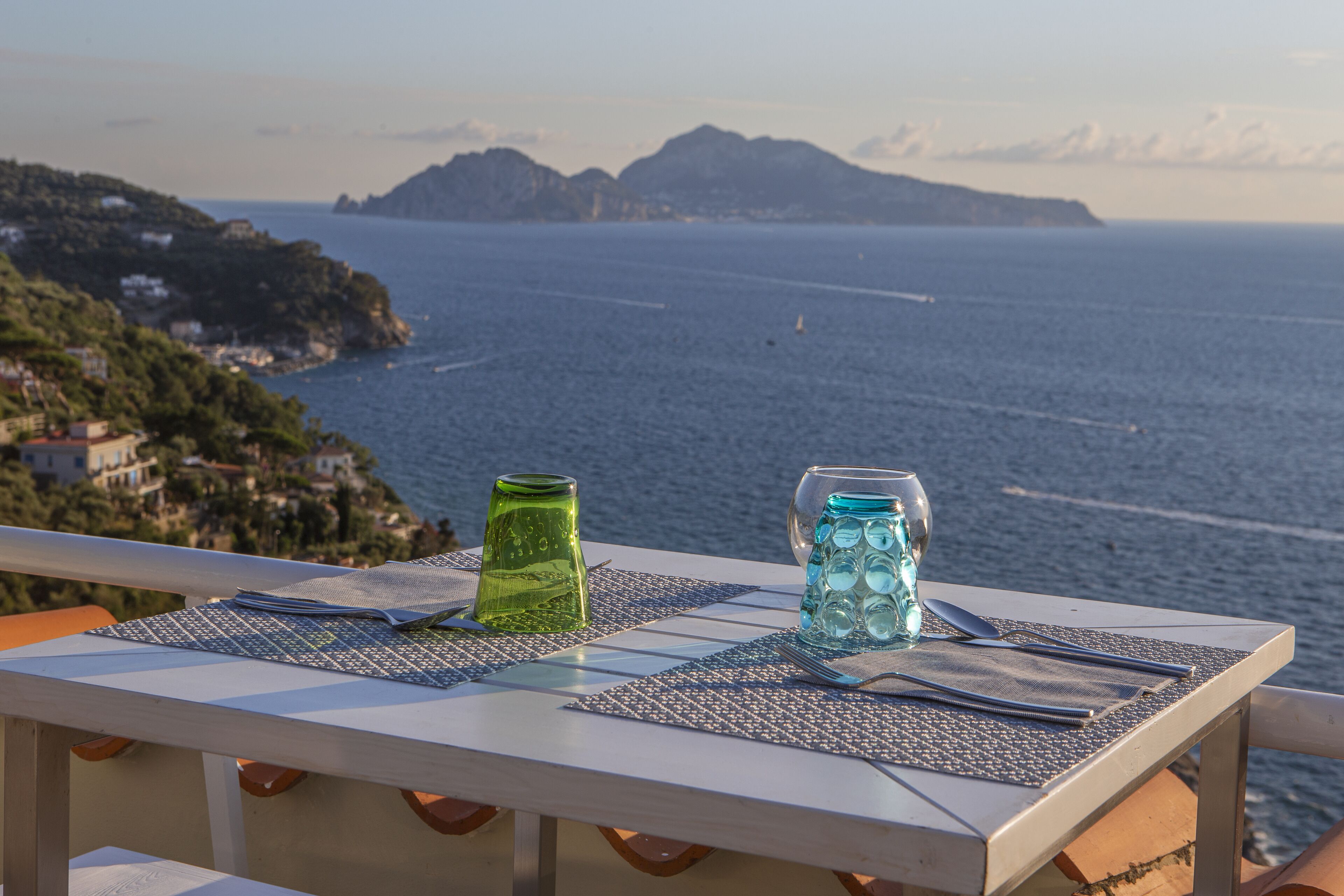 A table set with a green glass and a blue glass pitcher, overlooking a coastal view with mountains in the distance.
