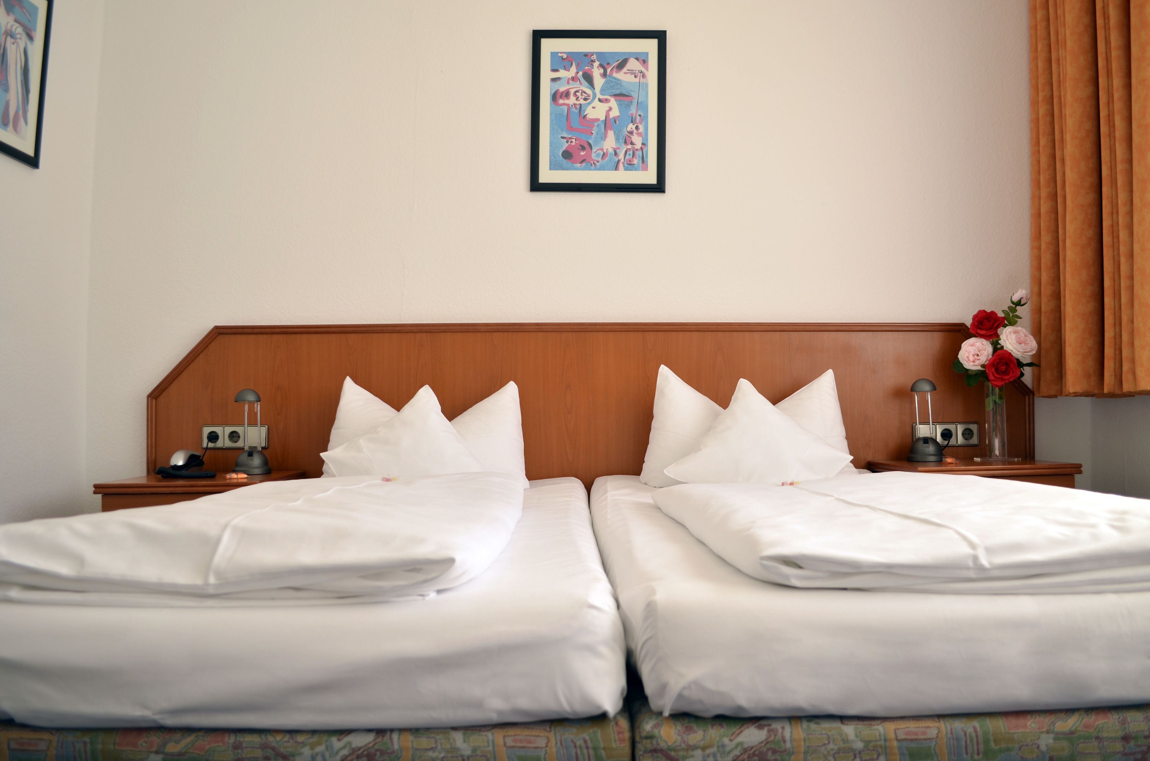 A double bed with white linens, a wooden headboard, a framed picture on the wall, and a vase with flowers on the nightstand.