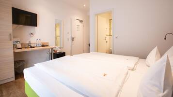 A hotel room with a bed, a desk with a chair, a TV mounted above the desk, and a bathroom visible through an open door.