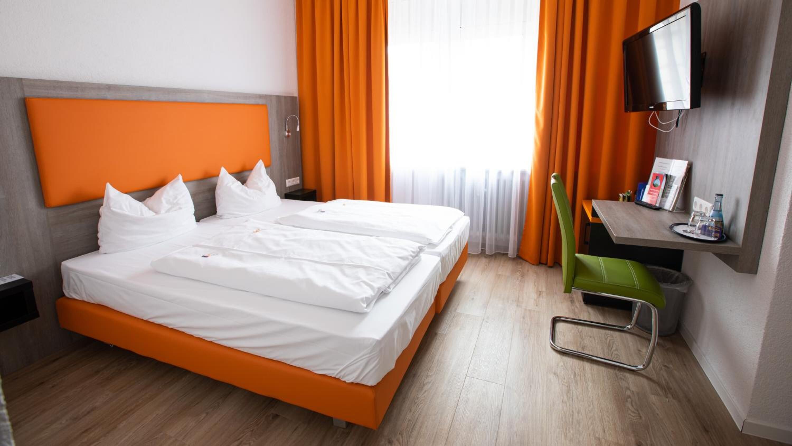 Quarto de hotel com duas camas, cortinas laranja, uma televisão e uma pequena mesa com objetos.