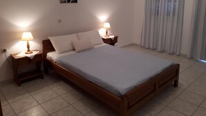 A bedroom with a wooden bed, two bedside tables with lamps, a painting on the wall, and a window with curtains. - Paradisos Oia (Santorini)