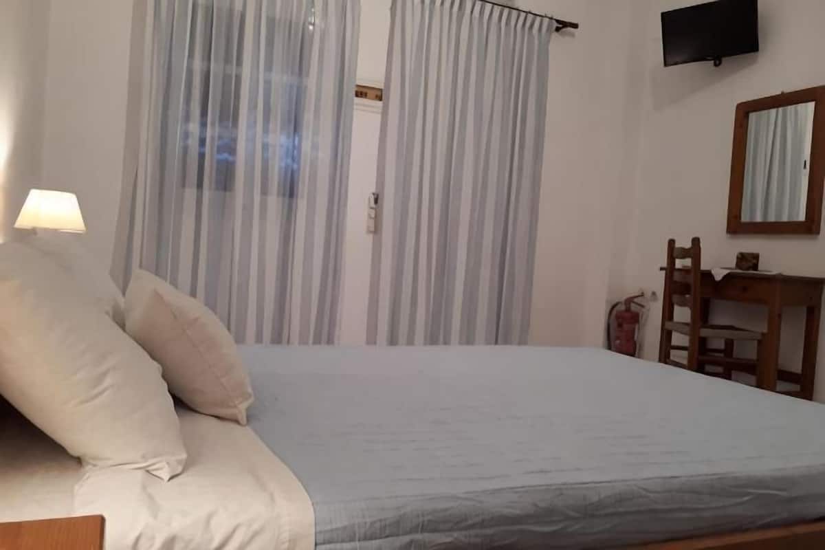 A bedroom with a bed, a desk, a chair, a TV, and a window with curtains.