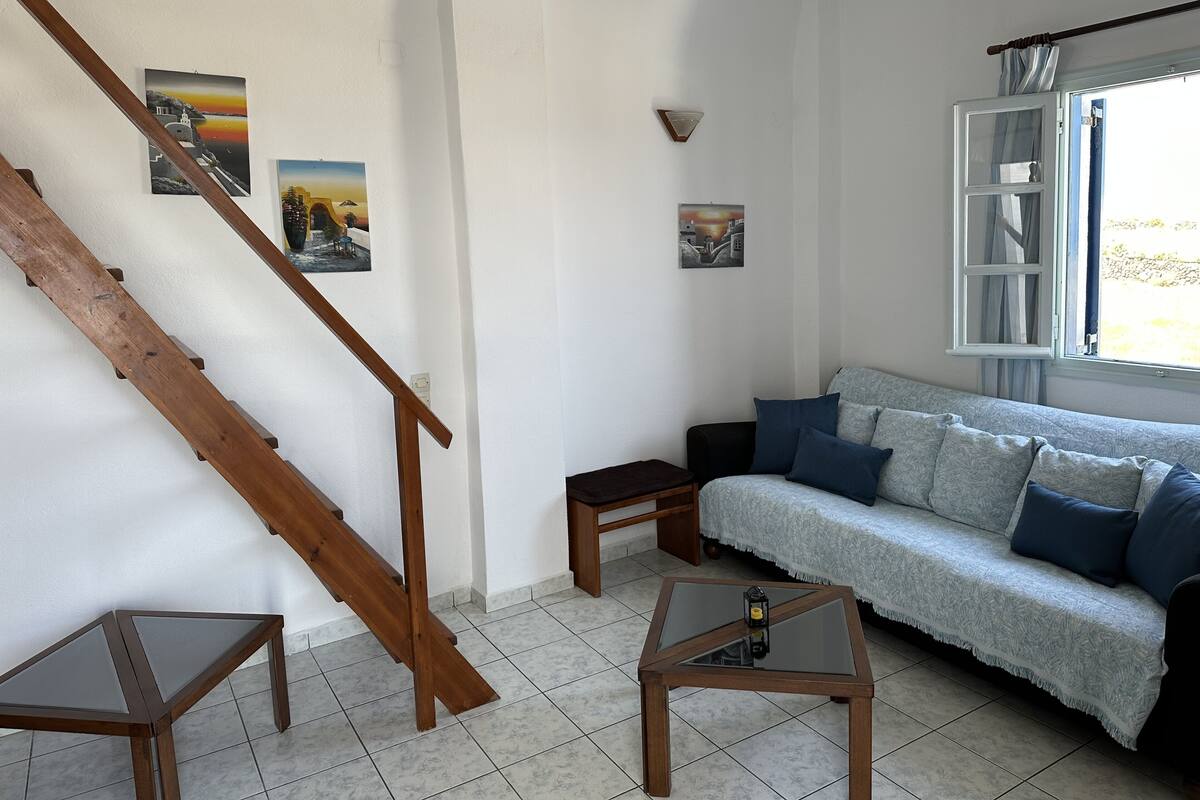A living room with a wooden staircase, a sofa with blue cushions, a small table with a lantern, and two wall-mounted pictures.